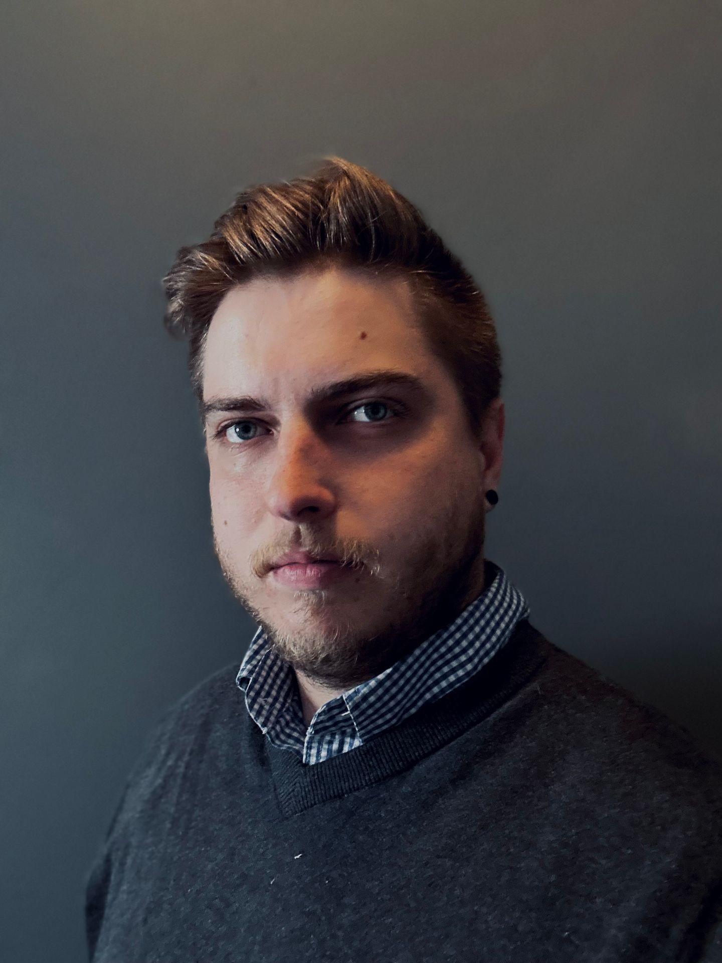 Person with short hair, wearing a checked shirt and dark sweater, standing against a gray background.